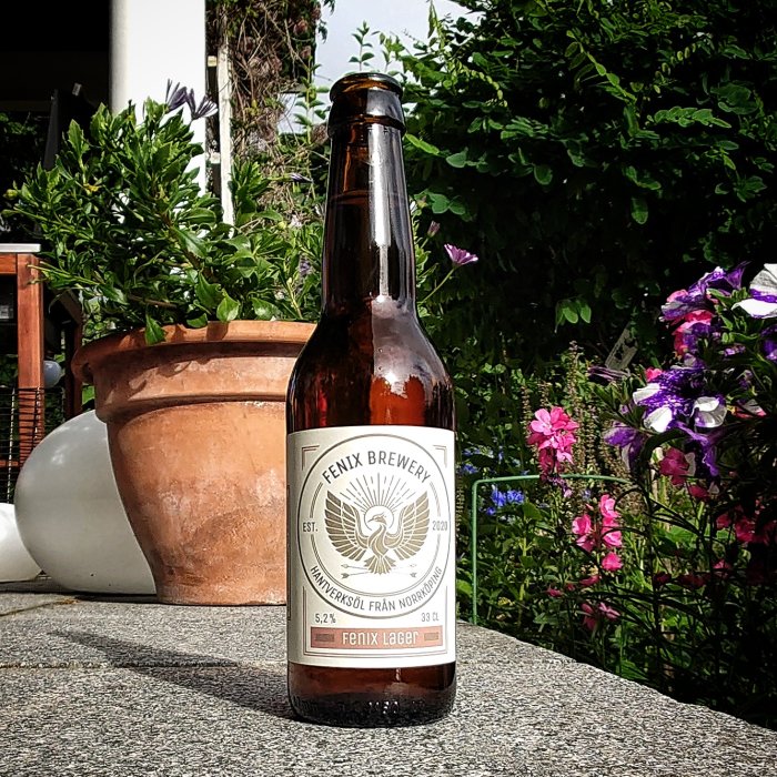En flaska Fenix Lager från Fenix Brewery står på en stenplatta med blommor och en kruka i bakgrunden.