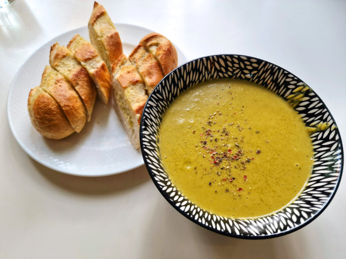 En tallrik purjolök- och potatissoppa bredvid vitlöksbröd på ett vitt bord.