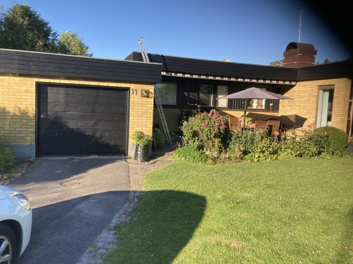 Ett gulmålat mexitegelhus med svarta träpartier, garageport och trädgårdsanläggning.