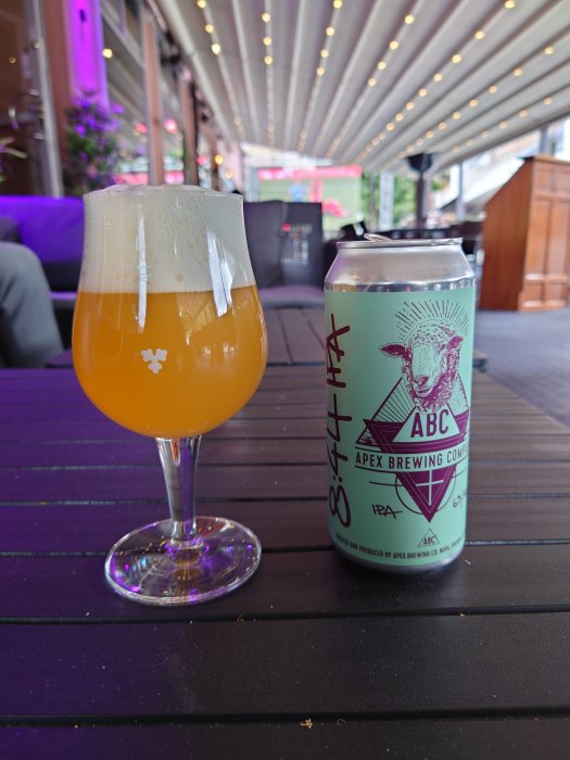 Ölglas med skummad öl bredvid en Apex Brewing IPA ölburk på ett bord på uteservering.