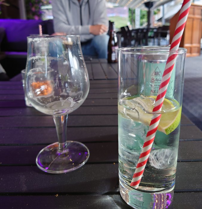 Drink med apple sour i högt glas med röd-vitrandigt sugrör, tom gin&tonic-glas i förgrunden, person i bakgrunden.