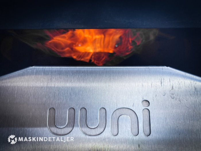 Närbild på en rostfri Uuni pizzaugn med eldflamman reflekterande i öppningen ovanför logotypen.