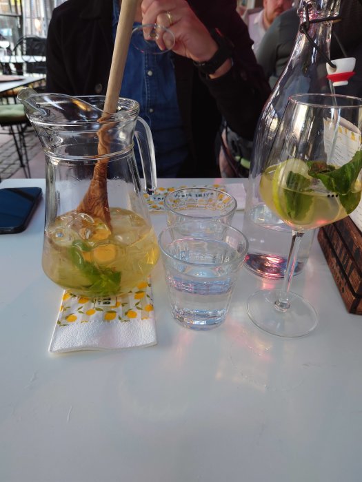 Karaff och glas med drinkar, fläder Basta Spritz och klar vätska, på ett bord med personer.