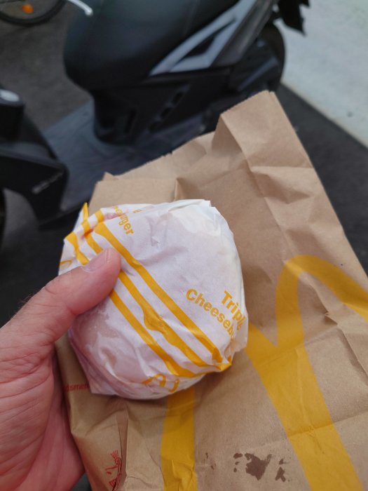 En hand håller en inslagen hamburgare märkt "Triple Cheese" med en papperspåse och en skoter i bakgrunden.