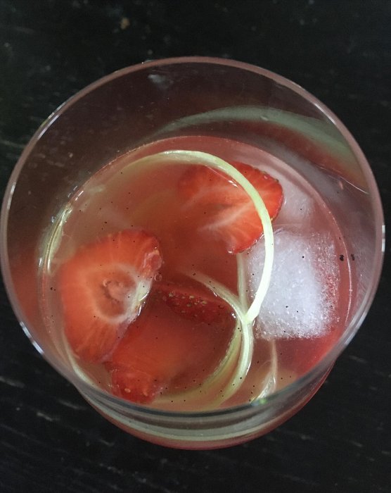 Ett glas rabarbersangria med skivade jordgubbar och citron på en svart bänkskiva.