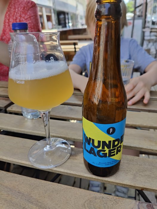 Belgisk IPA i glas bredvid tom ölflaska på ett utomhuscafébord, med suddig bakgrund.