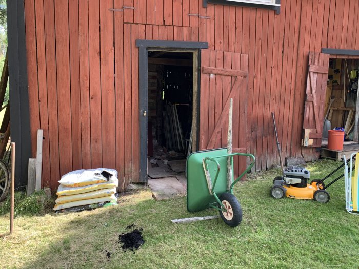 Grönt tomtrum med blommor intill en röd träbyggnad, en klippt gräsmatta, och trädgårdsredskap inklusive en röjsåg.