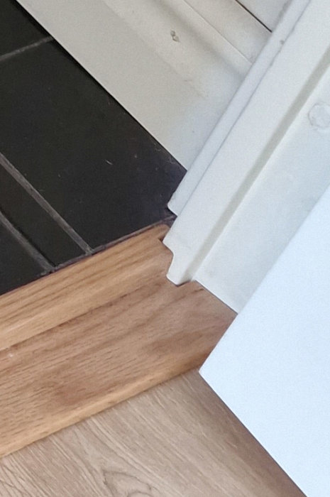 Tröskel mellan badrum med svart golv och rum med ljust trägolv, vit dörrpost.