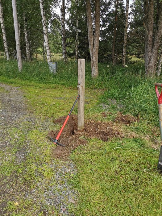 Grävarbete i trädgård med upptagen jord runt en borttagen grindstolpe och en spade lutad mot stolpen.