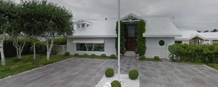 Modern villa med vita väggar, grått tak, stenlagd uppfart och gröna klätterväxter.