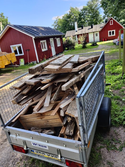 Släpvagn full med rivet trä och plankor, med röda stugor i bakgrunden.