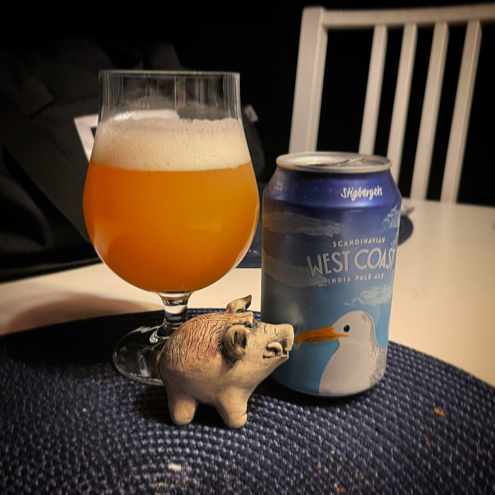 Ölglas med skum på en bricka intill en West Coast IPA-ölburk med ett vildsvinsfigurin.