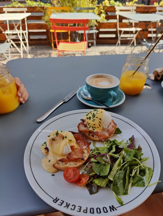 Tallrik med två Eggs Benedict och sallad på ett cafébord, juice och cappuccino i bakgrunden.