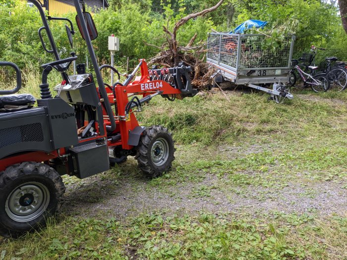 Minigrävare med gafflar lyfter en grenbunt vid en trailer och cyklar på en gräsmark.