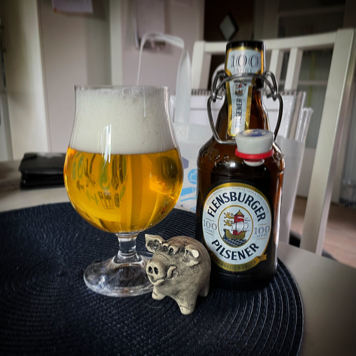 Ölglas fullt med öl intill en stängd Flensburger Pilsener-flaska och en liten grisknorr-figur.