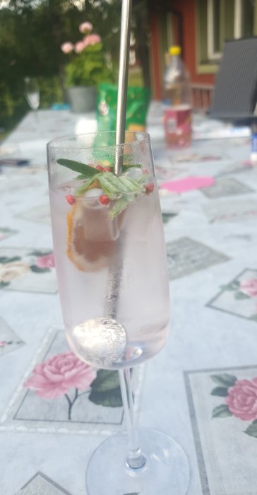 Drinkglas med en GT på ett blommönstrat bord, grönt garnering och iskuber, oskarp bakgrund.