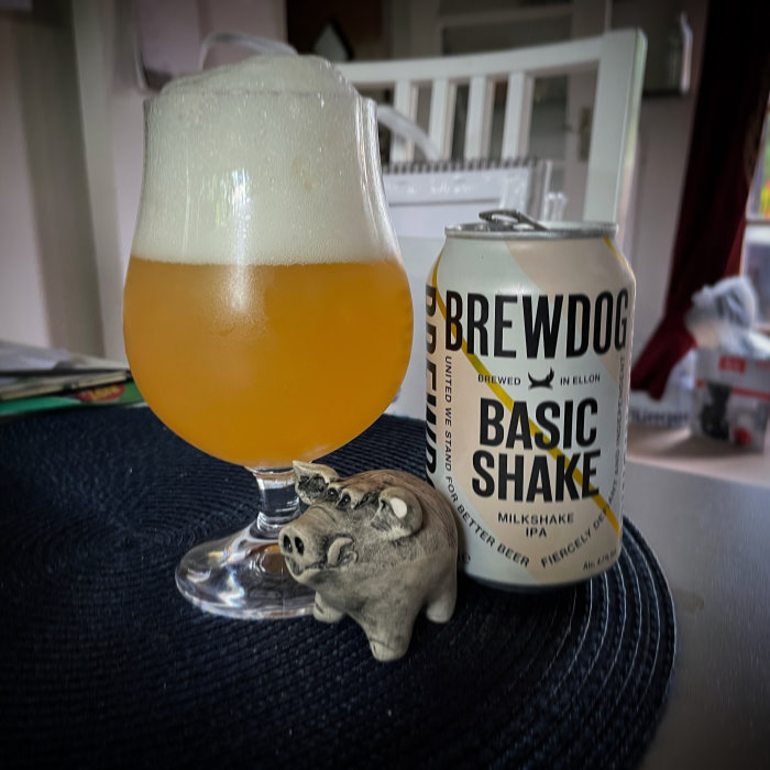 Ölglas med skummad öl och Brewdog Basic Shake burk bredvid en metallgris på bord.