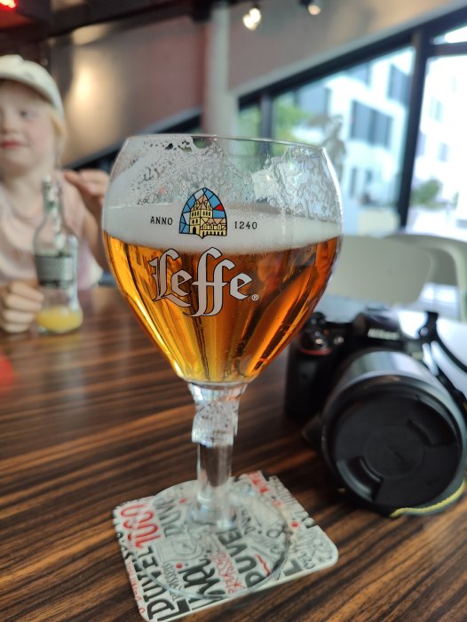 Ett glas Leffe-öl med skum på kanten, på en coaster, med en kamera och en suddig person i bakgrunden.