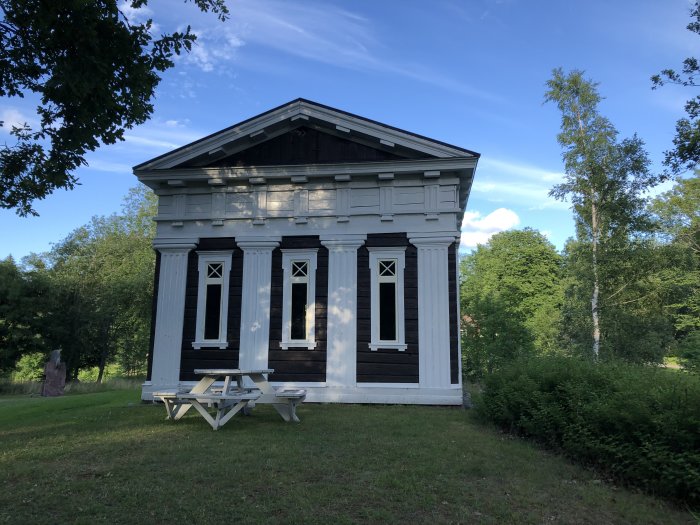 En klassisk träbyggnad med vita pelare och fönsterluckor, samt en picknickbänk framför.