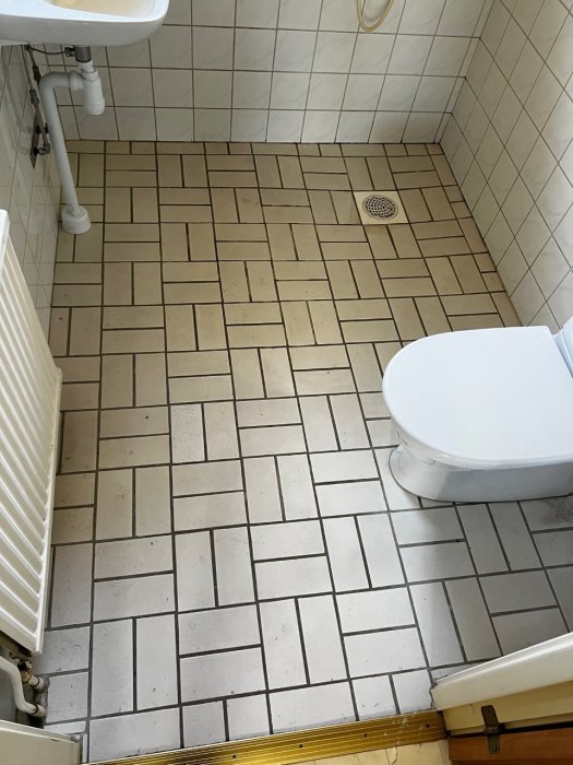 Före-bild av badrummet före renovering med kaklade väggar, golv med kakelplattor, toalettstol och radiator synliga.