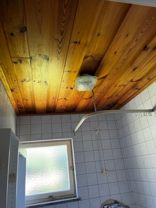 Före bild av badrum med träbeklädd tak, kaklade väggar, fönster med frostat glas och demonterat duschmunstycke.