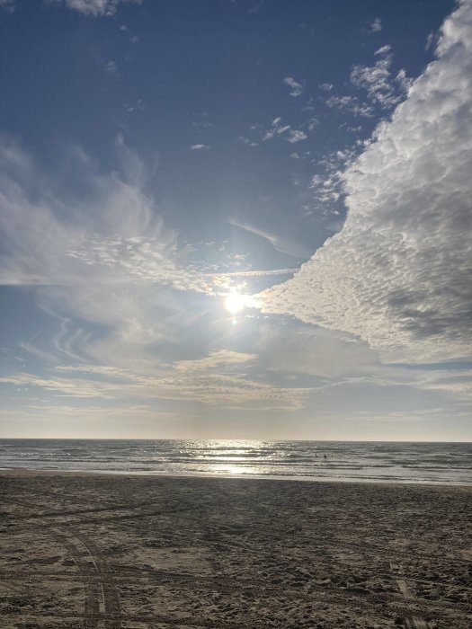 Strand med fotspår i sanden, skimrande hav och solen som delvis döljs av dramatiska moln på himlen.