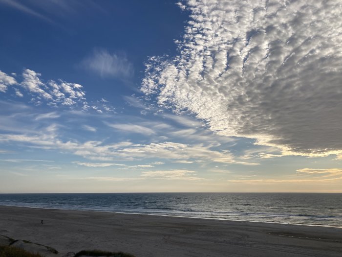 Vidsträckt sandstrand med lugnt hav och himmel med blandade molnformationer vid solnedgång.