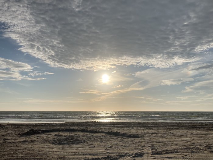 Solnedgång över havet med vågor och moln på himlen, sedd från en sandig strand med spår av däck.