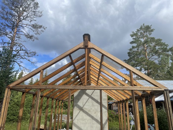 Trästomme för ett byggnadsverk med takstolar och glaspartier under molnig himmel.