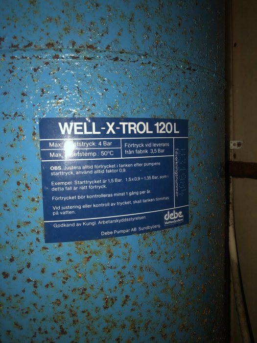 Rostig blå trycktank med WELL-X-TROL-etikett i en källare, tecken på korrosion och åldrande.