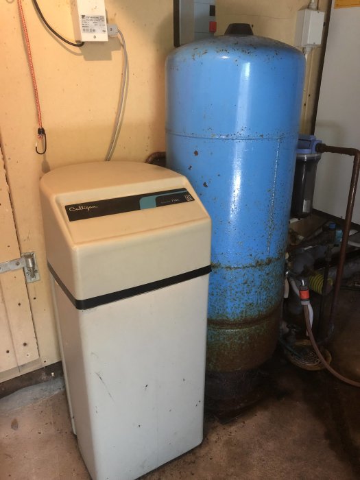 Vattenreningsanläggning i källare med en vit mjukvattenenhet bredvid en blå trycktank som visar tecken på rost och slitage.