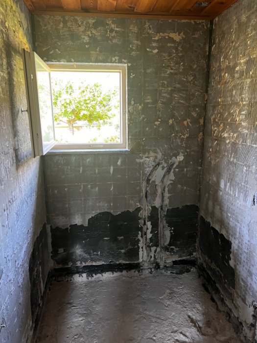 Ett badrum under renovering med avbilade väggar och golv, exponerat avloppsrör och fönster mot grönska.