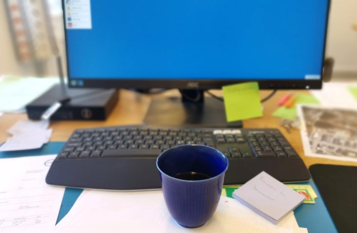 En blå mugg med kaffe framför en suddig arbetsstation och datorskärm.