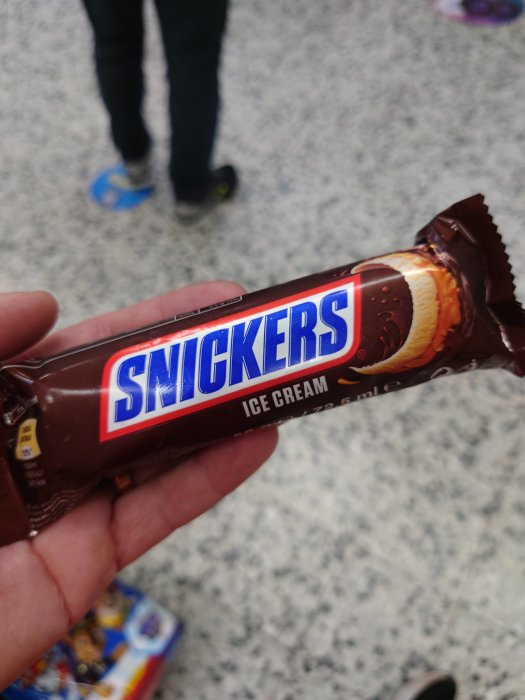 Hand håller en Snickers glasspinne framför suddig bakgrund av golv och personers fötter.