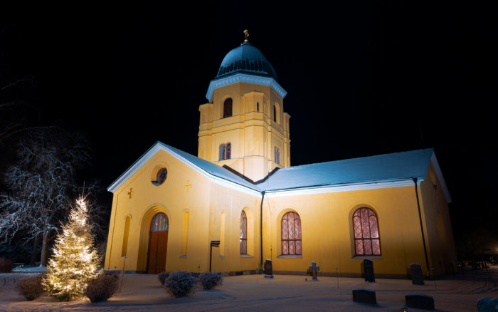 En välbelyst kyrka på natten med strålkastare och en julgran i snöig miljö.