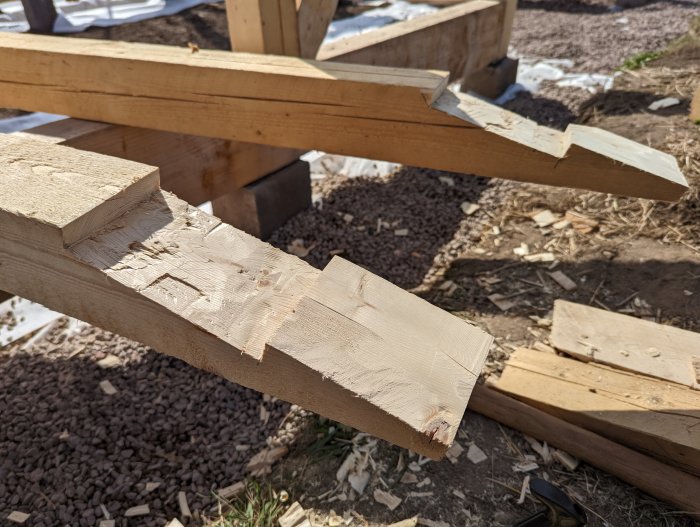 Trähantverk av takåsar under konstruktion i en byggarbetsplats med synlig grus och spillvirke i bakgrunden.