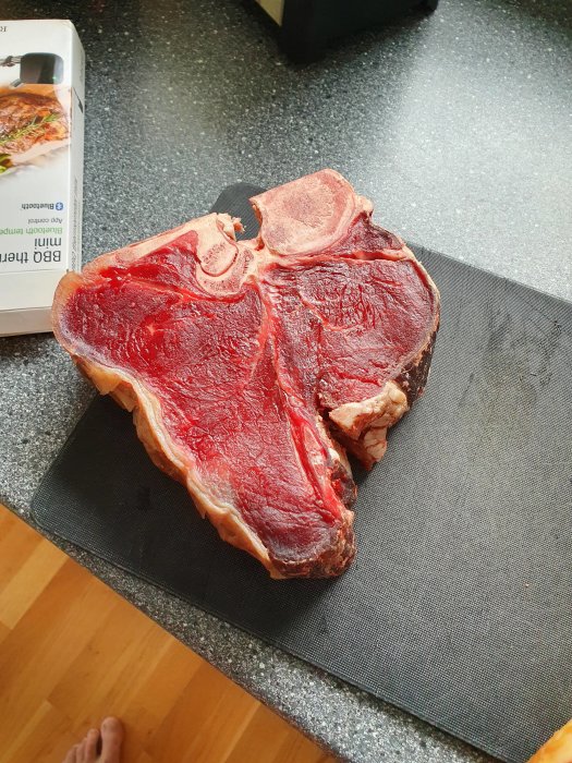 Ett stort T-benstek på en skärbräda i köket, fotograferat ovanifrån med en fot synlig.