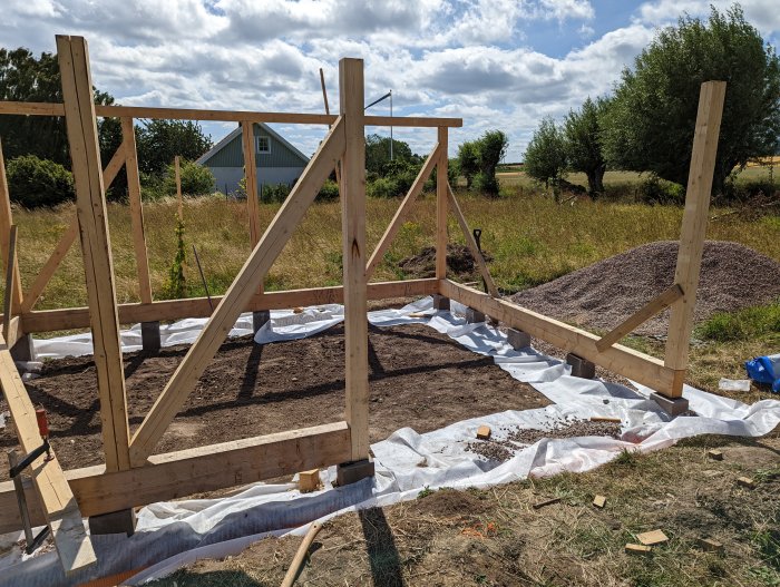 Pågående bygge av träkonstruktion med färdiga långsidor, uppsatt takås, dränering och utskridat grus på markduk.