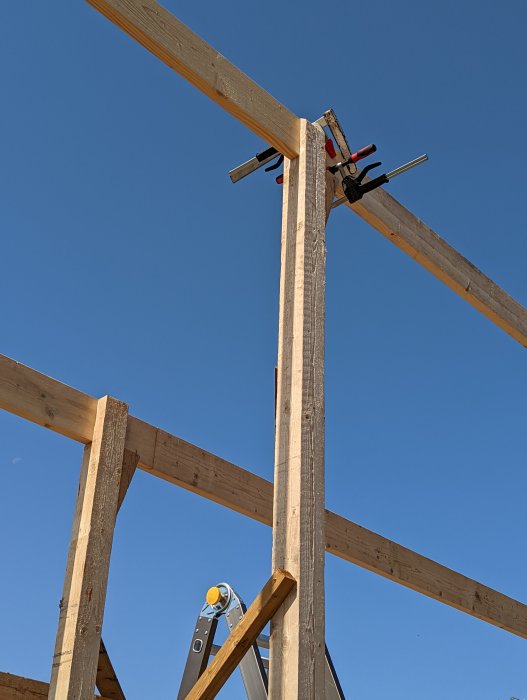 Träkonstruktion med åsar som möts vid stolpe, fixerade med klämmor, mot en klarblå himmel.