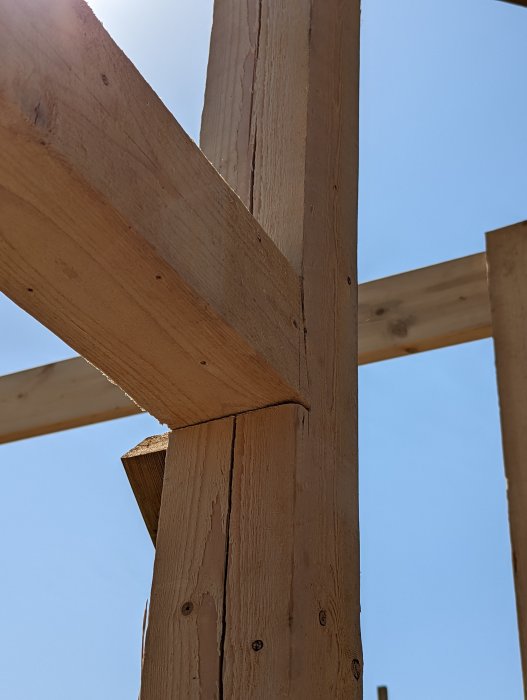 Anslutning mellan träås och stolpe med urtag i byggprojekt mot en klarblå himmel.