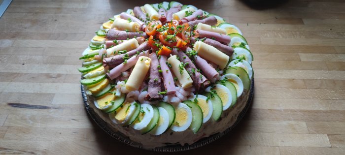Hemmagjord smörgåstårta med köttpålägg, ost, ägg, räkor, gurka och tomat på en träyta.