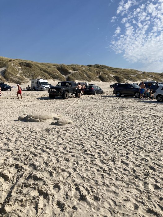 Bilar parkerade på en sandig strand med människor och sandfigurer i förgrunden.