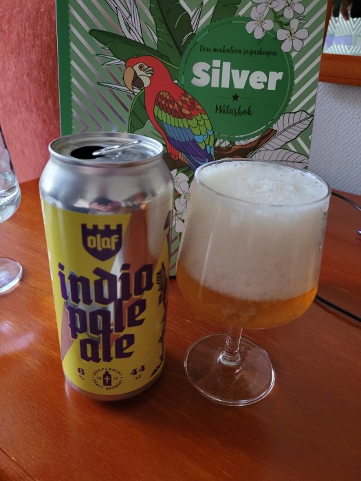 Burk finsk IPA med texten "India Pale Ale" bredvid ett glas öl med skumkrona, på ett bord med målarbok i bakgrunden.