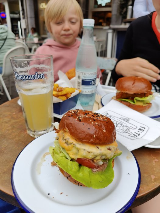 En halvdrucken veteöl i glas, hamburgare på tallrik, vattenflaska och en person i bakgrunden.