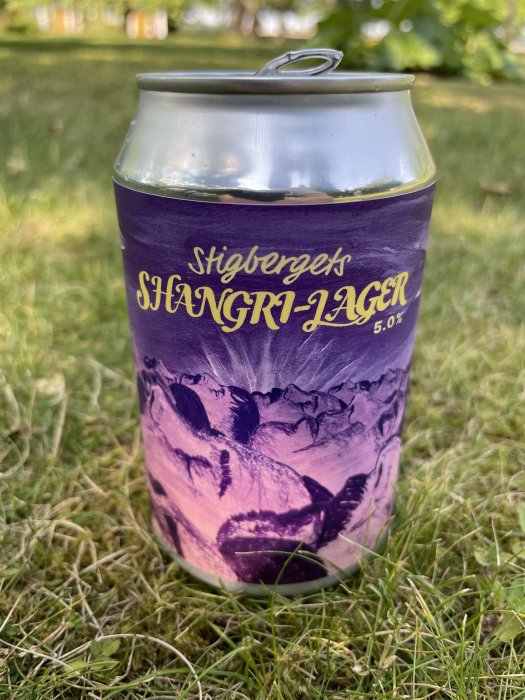 Öppnad ljust lila ölburk märkt "Shangri-Lager" i gräset, svalkande dryck för en varm dag.