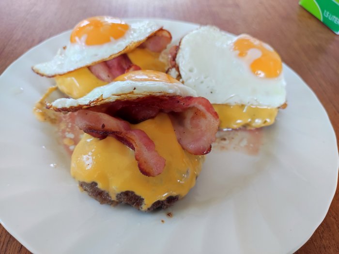 Lammfärsbiffar toppade med smält ost, krispiga baconskivor och stekta ägg på en vit tallrik.