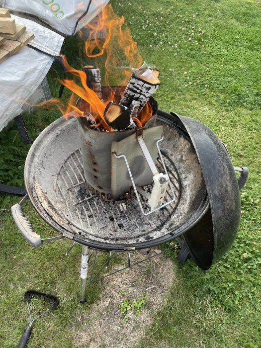 Klotgrill med igångsatta kol i en startchimney, flammor och gräsmatta i bakgrunden.