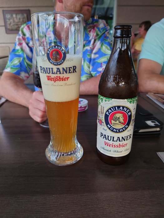 Paulaner Weissbier i ett glas bredvid öppnad flaska på bord med delvis synlig person i bakgrunden.