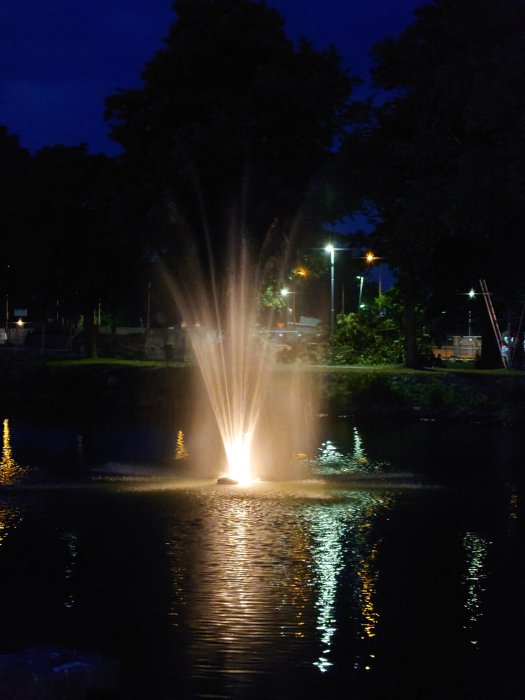 Belyst fontän med vattenstrålar i en damm under kvällstid.
