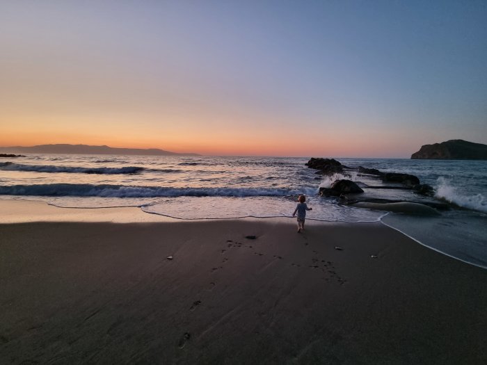 Barn leker på stranden vid solnedgång med vågor och klippor i bakgrunden